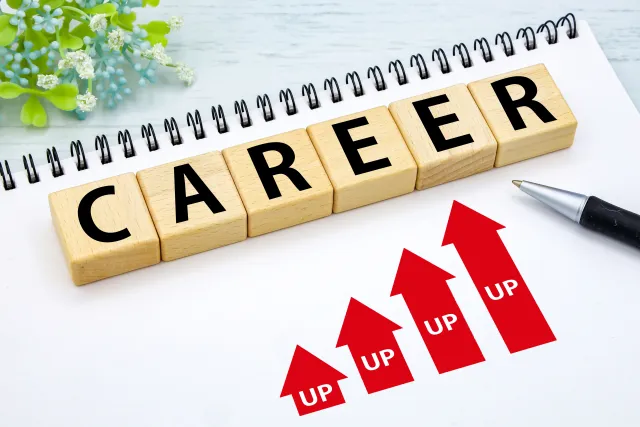 「CAREER」の文字を並べた木製ブロックと、上向きの「UP」の矢印が描かれたノート